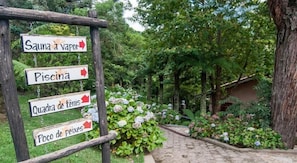 A wooden signpost with directional arrows pointing to various amenities: Sauna, Piscina, Quadra de tenis, and Poço de peixes. - Pousada Ricanto Amore Mio (Camanducaia)