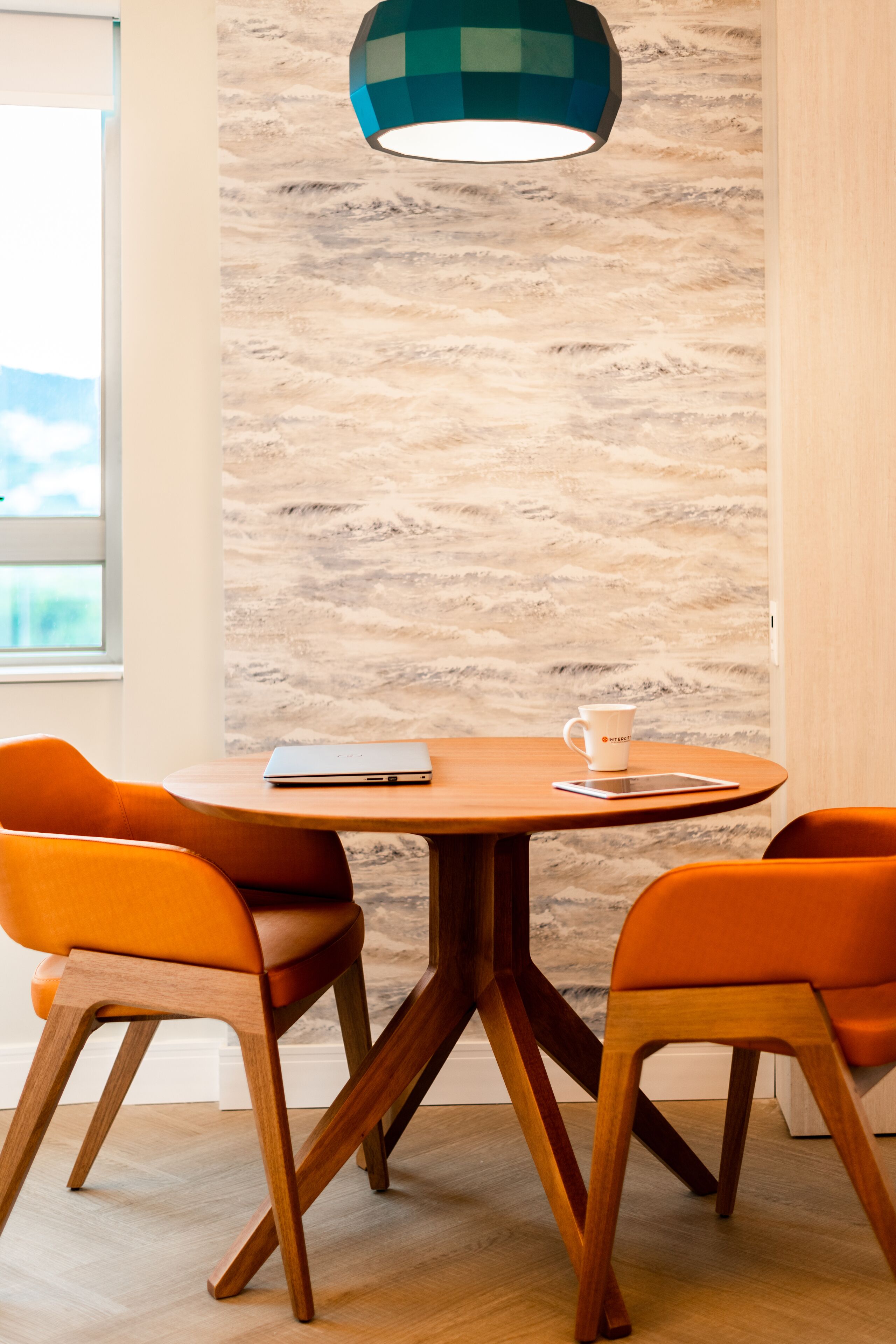 A dining area with a round wooden table, two orange chairs, a tablet, and a white mug on the table.