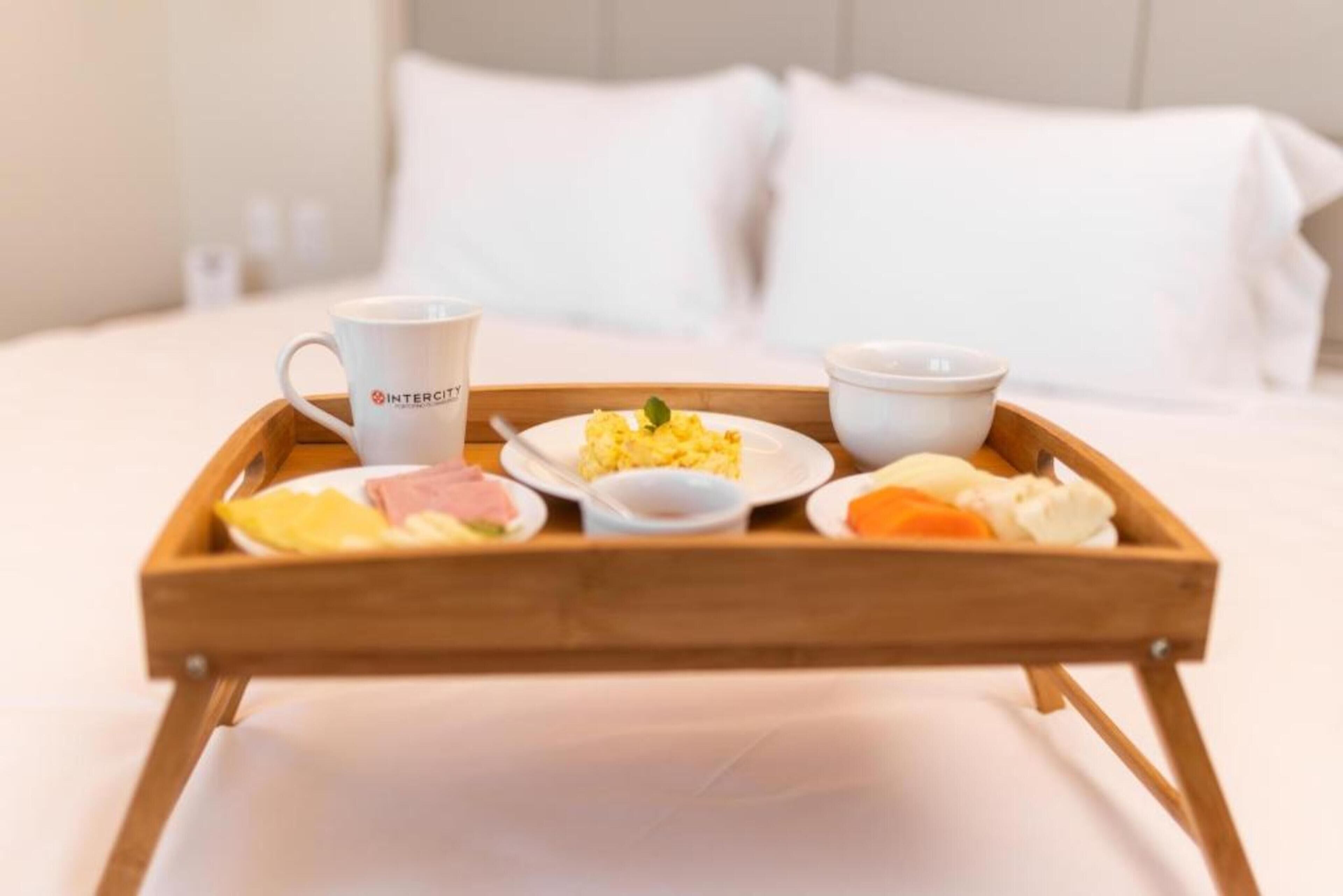 A wooden tray with breakfast items including scrambled eggs, ham, and toast, placed on a bed with white bedding and pillows.