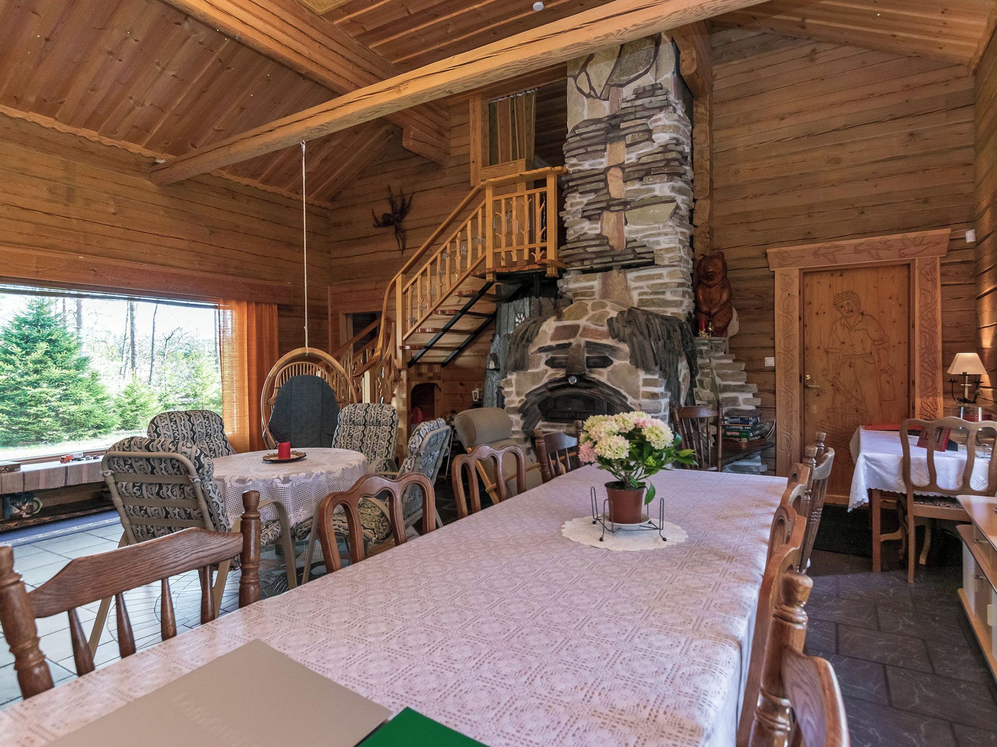A wooden interior with a stone fireplace, dining area with chairs and tables, and a staircase leading to an upper level.