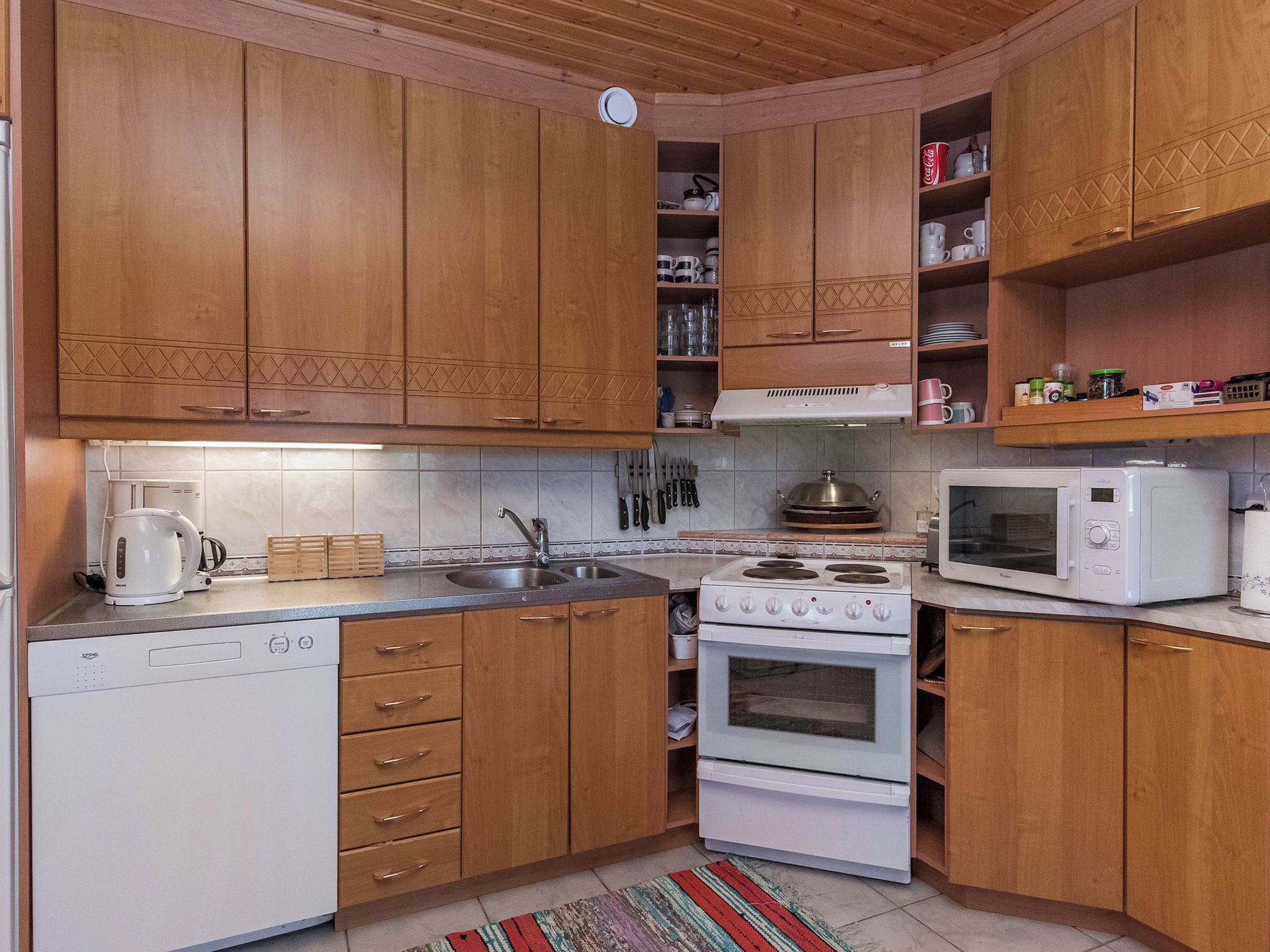 Een keuken met houten kastjes, een witte vaatwasser, een fornuis, een magnetron en een spoelbak.