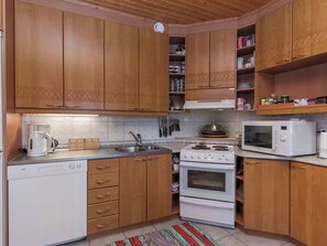 A kitchen with wooden cabinets, a white dishwasher, a stove, a microwave, and a sink. - Villa kesäranta by Interhome (Kesälahti)