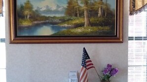 Een kamer met een gekaderd landschap, een kleine Amerikaanse vlag en een vaas met paarse bloemen op een houten tafel.