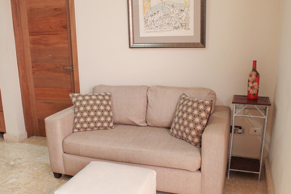 A beige sofa with patterned cushions, a small ottoman, a framed artwork on the wall, and a bottle on a metal side table.