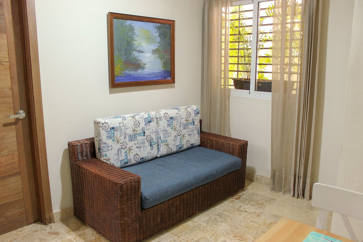 A wicker sofa with patterned cushions, a painting of a landscape, and a window with curtains.