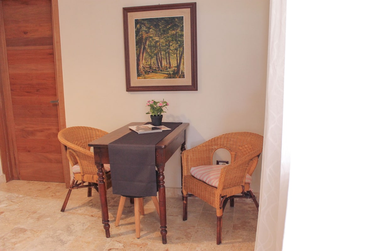 A dining area with a wooden table, two wicker chairs, a framed painting on the wall, and a potted plant.