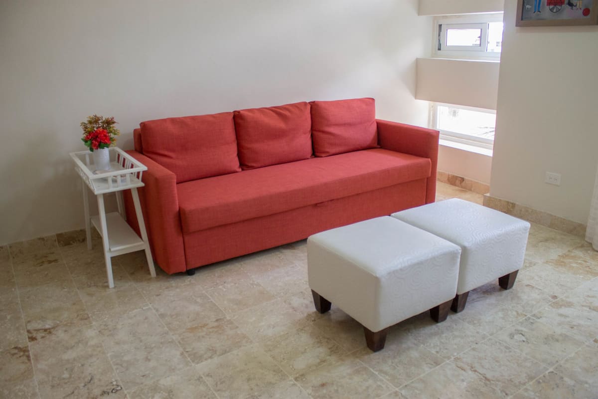 A red sofa, a white ottoman, and a small white side table with a vase of flowers in a room with tiled flooring and a window.
