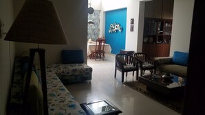 Una sala de estar con un sofá, sillas, una mesa de centro y una lámpara. Se observa un comedor al fondo, con una mesa y sillas.