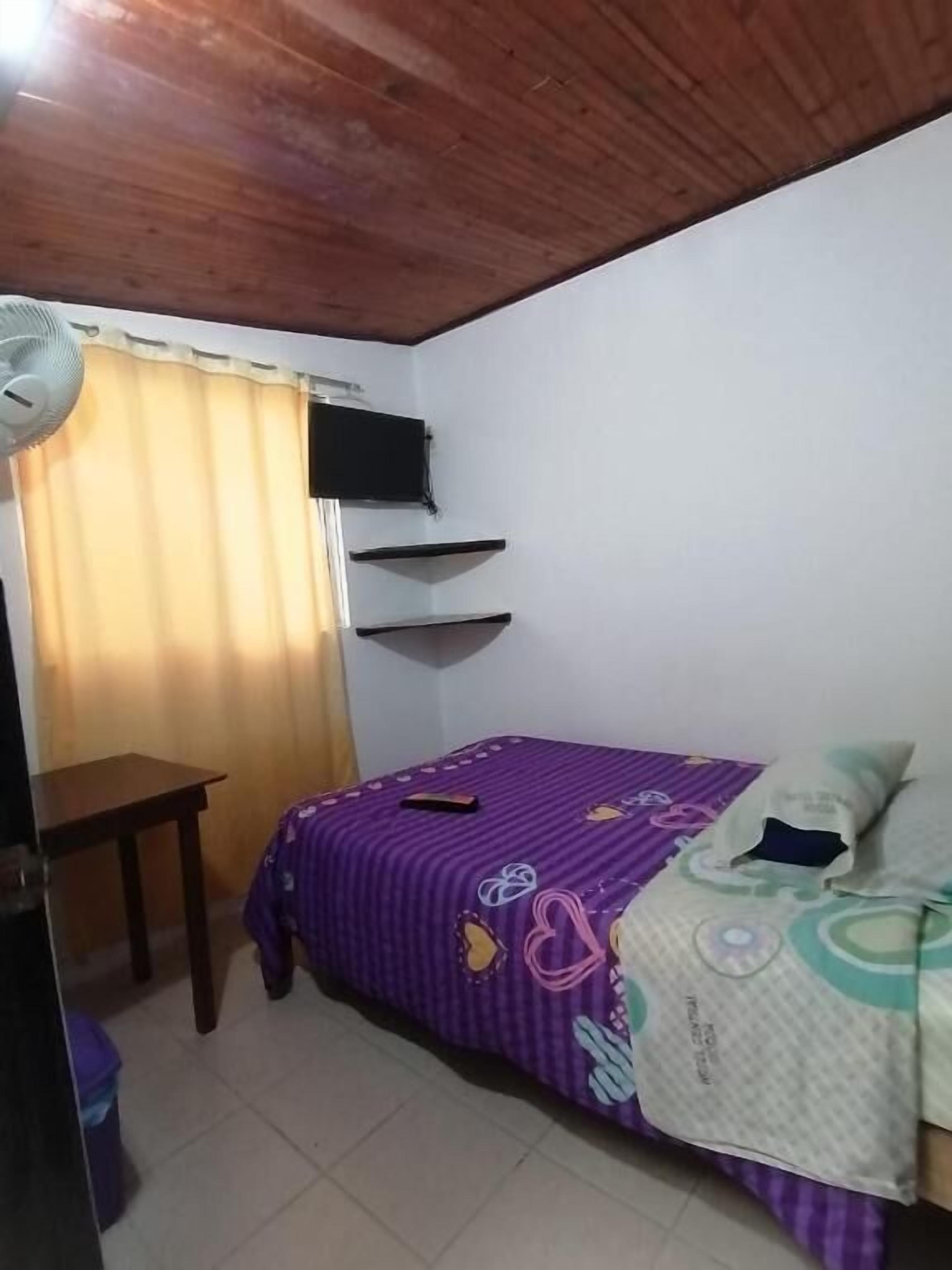 A small bedroom with a bed, a desk, a chair, a TV mounted on the wall, and a window with curtains.