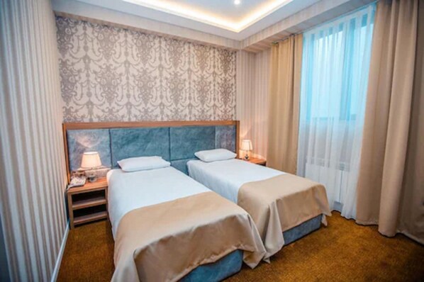 A hotel room with two beds, a patterned wallpaper, a large window with curtains, and a nightstand with a lamp.