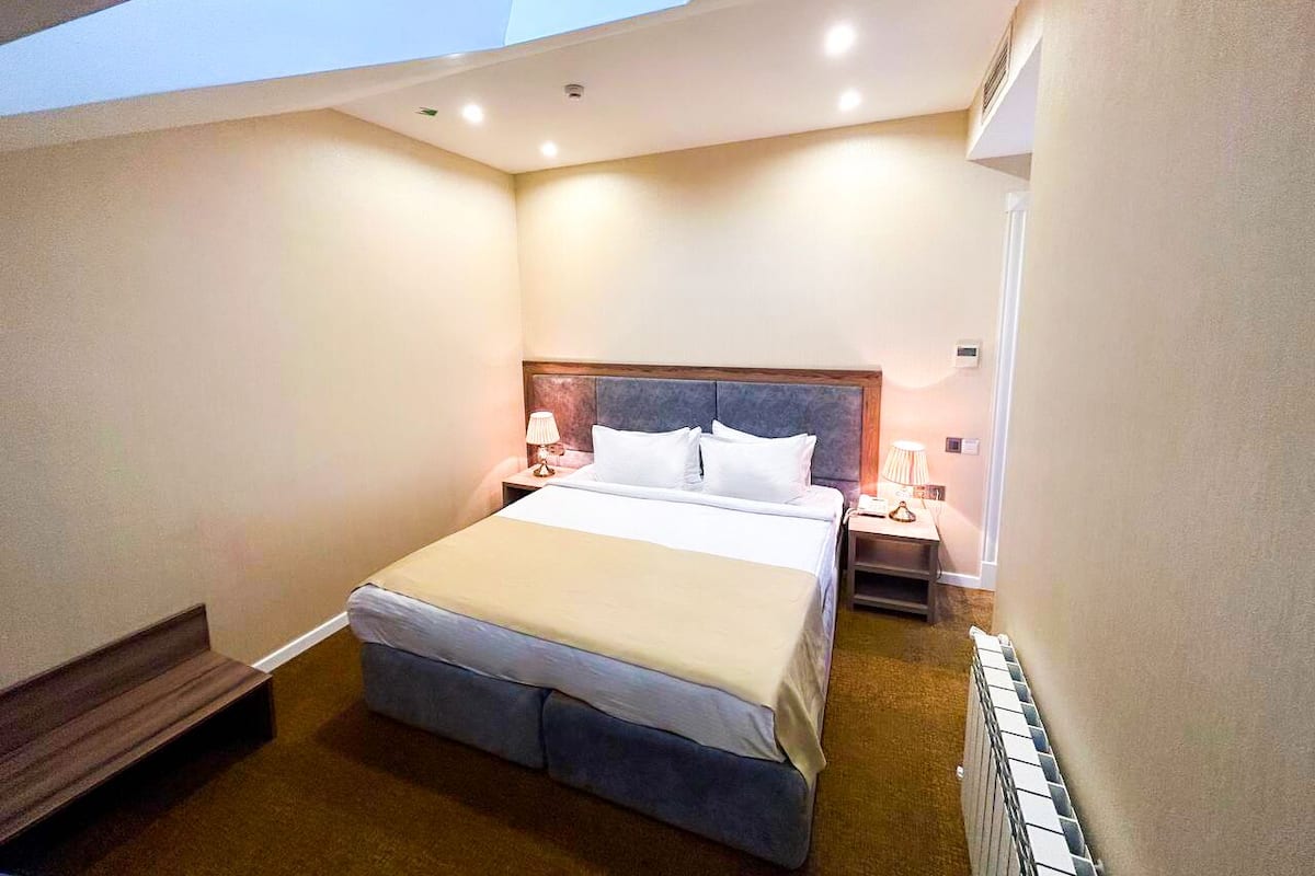 Une chambre d’hôtel avec une lucarne, un lit recouvert de draps blancs, une table de chevet avec une lampe et un banc en bois.