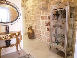 Una habitación con pared de piedra, una consola de madera con un florero, un espejo y un aparador de vidrio con objetos decorativos.