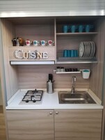 Cocina compacta con estufa a gas, un lavamanos y alacenas de madera. Se observan repisas con adornos y utensilios de cocina.