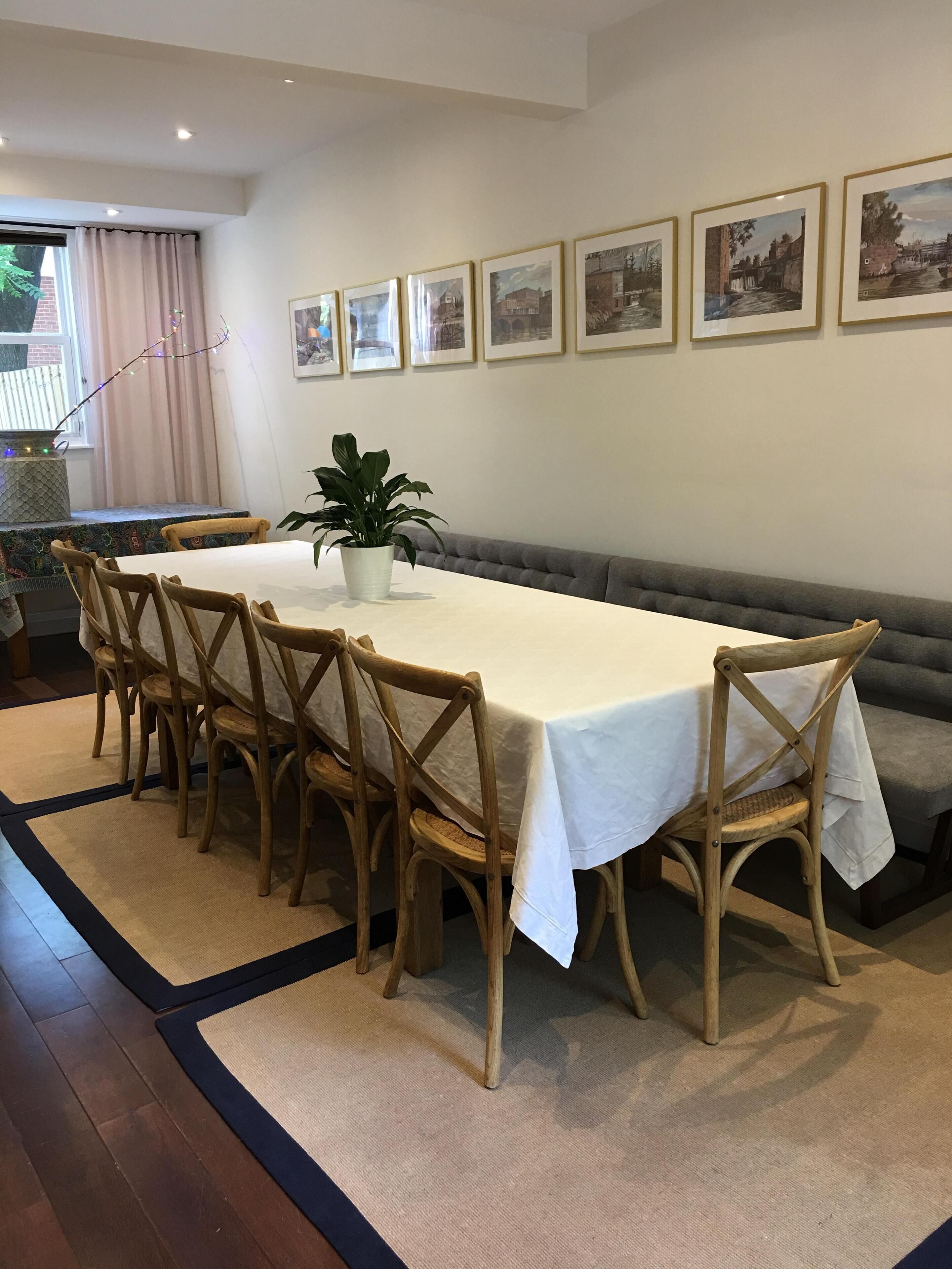 A dining area with a long table, six wooden chairs, a white tablecloth, a potted plant, and a sofa.