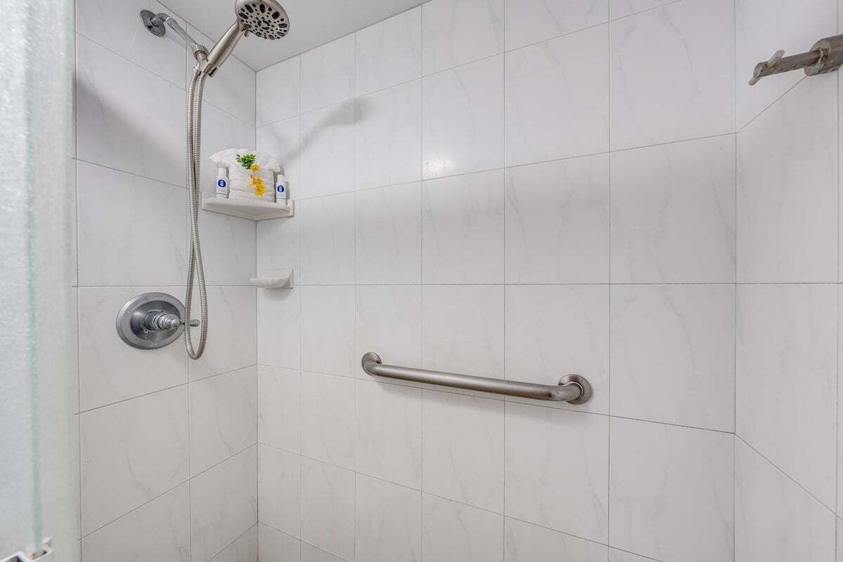 Un baño con baldosas blancas, un cabezal de ducha manual, un asidero y un estante con artículos de aseo.