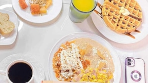 Uma pessoa lendo um livro à mesa, com itens de café da manhã como waffles, torradas e uma xícara de café.