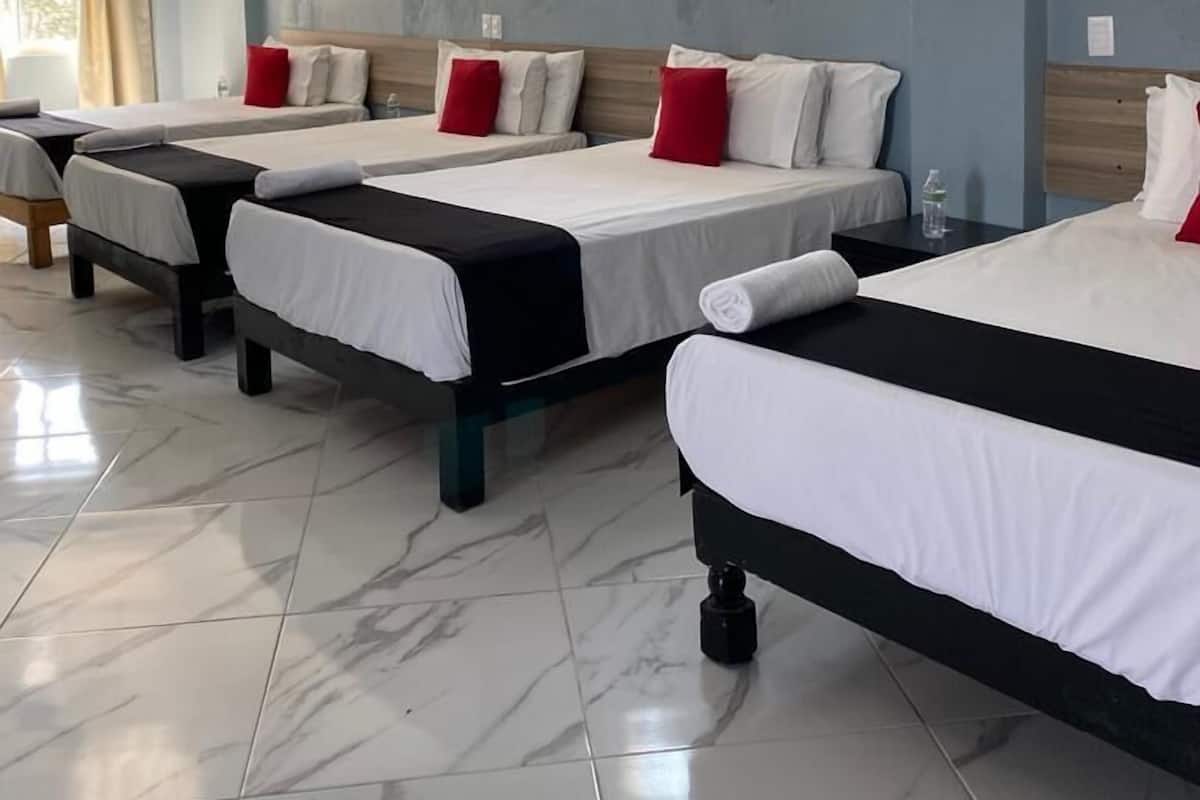 A hotel room with four single beds, each with white bedding and red pillows. There is a large window providing natural light, and an air conditioning unit mounted on the wall.
