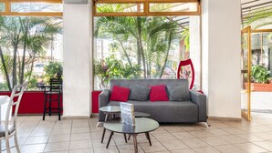 Um interior moderno com um sofá cinzento, almofadas vermelhas, uma mesa de centro de vidro e vista para palmeiras através de grandes janelas.