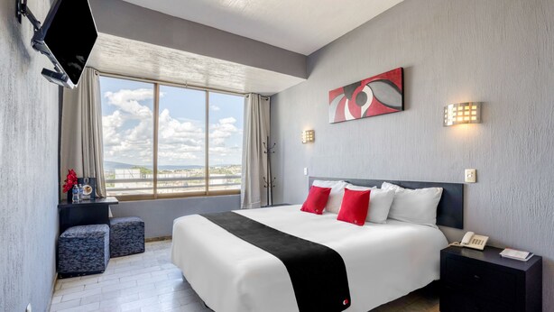 Quarto de hotel moderno com uma cama grande, televisão de tela plana na parede, mesa de cabeceira com telefone e janela com vista para a cidade.