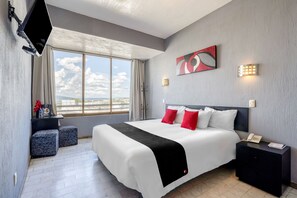 A modern hotel room with a large bed, a flat-screen TV mounted on the wall, a bedside table with a phone, and a window with a view of the cityscape.