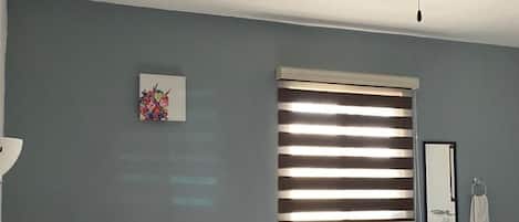 A bedroom with a bed, pillows, a ceiling fan, a window with blinds, a wall-mounted mirror, and a small artwork.