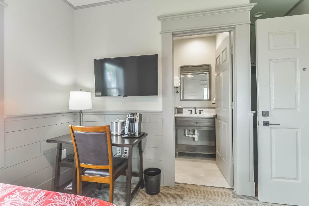 A hotel room with a bed, a desk with a chair, a television, a small table with a lamp, a trash bin, and an open door leading to a bathroom.