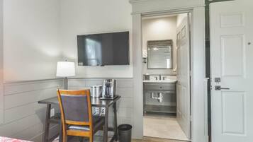 Habitación de hotel con cama, escritorio y silla, televisor, mesita de noche con lámpara, cesto de basura y una puerta abierta que da a un baño.