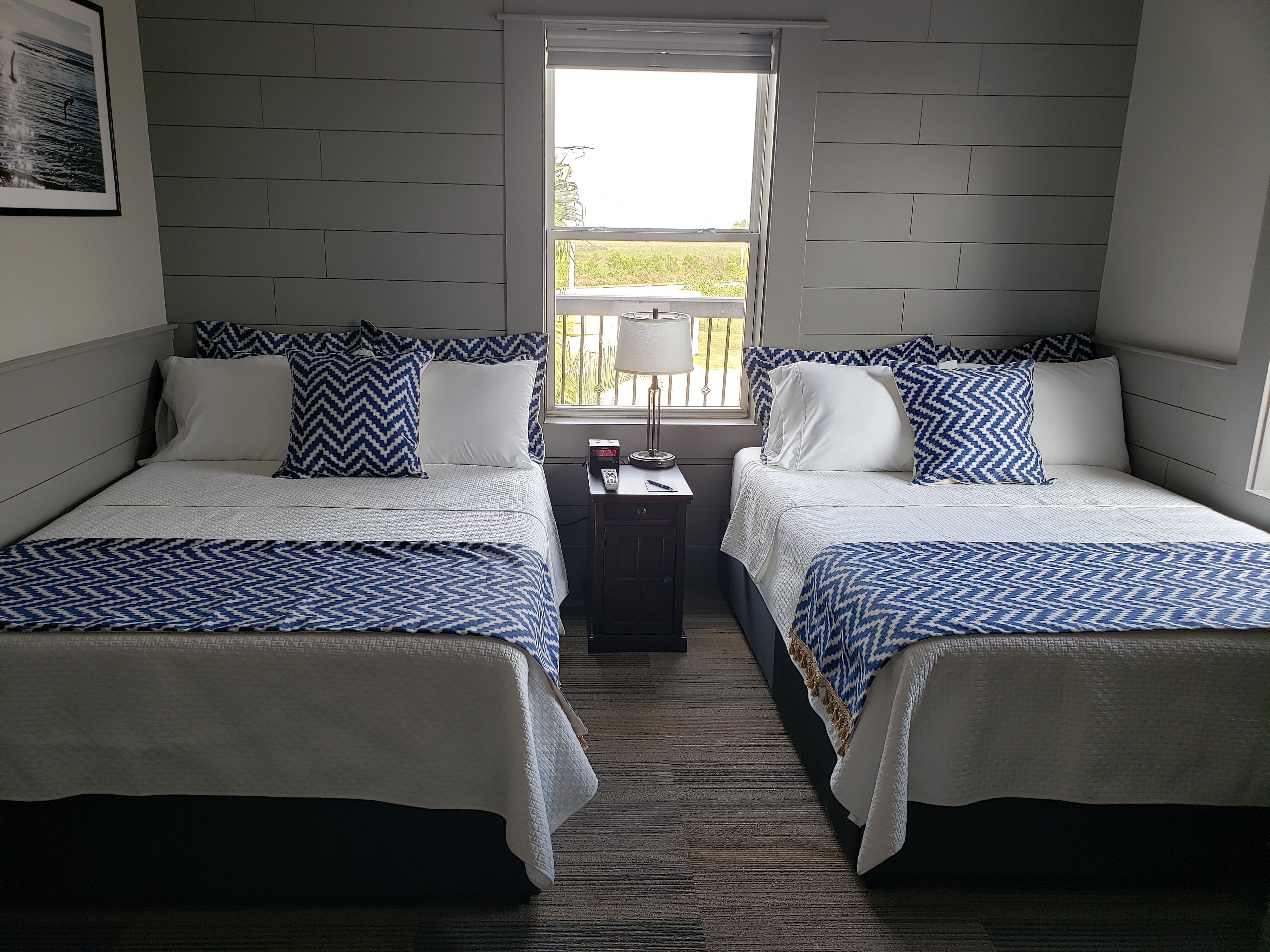 A room with two beds, each with a patterned bedspread, a nightstand with a lamp, and a window with a view of greenery.