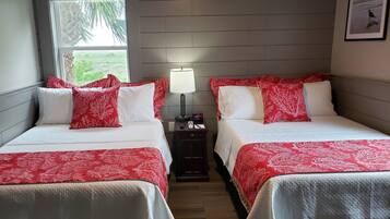 A hotel room with two beds, each with red and white bedding, a nightstand with a lamp, and a window with a view of greenery.