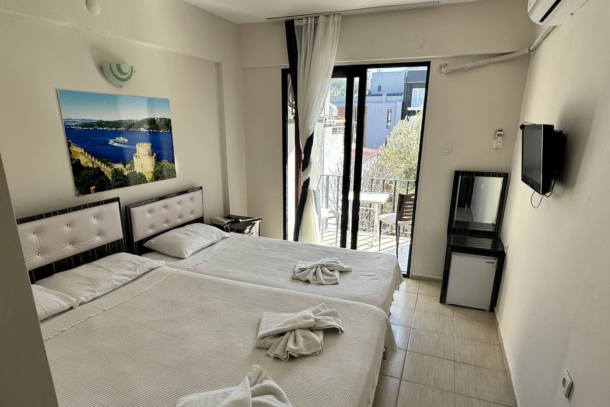 A hotel room with a bed, a painting, a TV, and a balcony with a view of buildings.