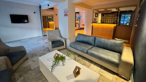 A modern living room with a grey sofa, two armchairs, and a white coffee table with a plant. A TV is mounted on the wall, and a reception desk is visible in the background. - Erciyes Otel Kusadasi (Kusadasi)