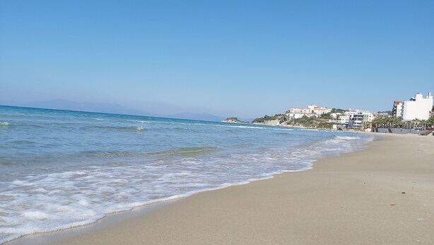 A beach with clear blue water, a sandy shore, and buildings in the background. - Erciyes Otel Kusadasi (Kusadasi)