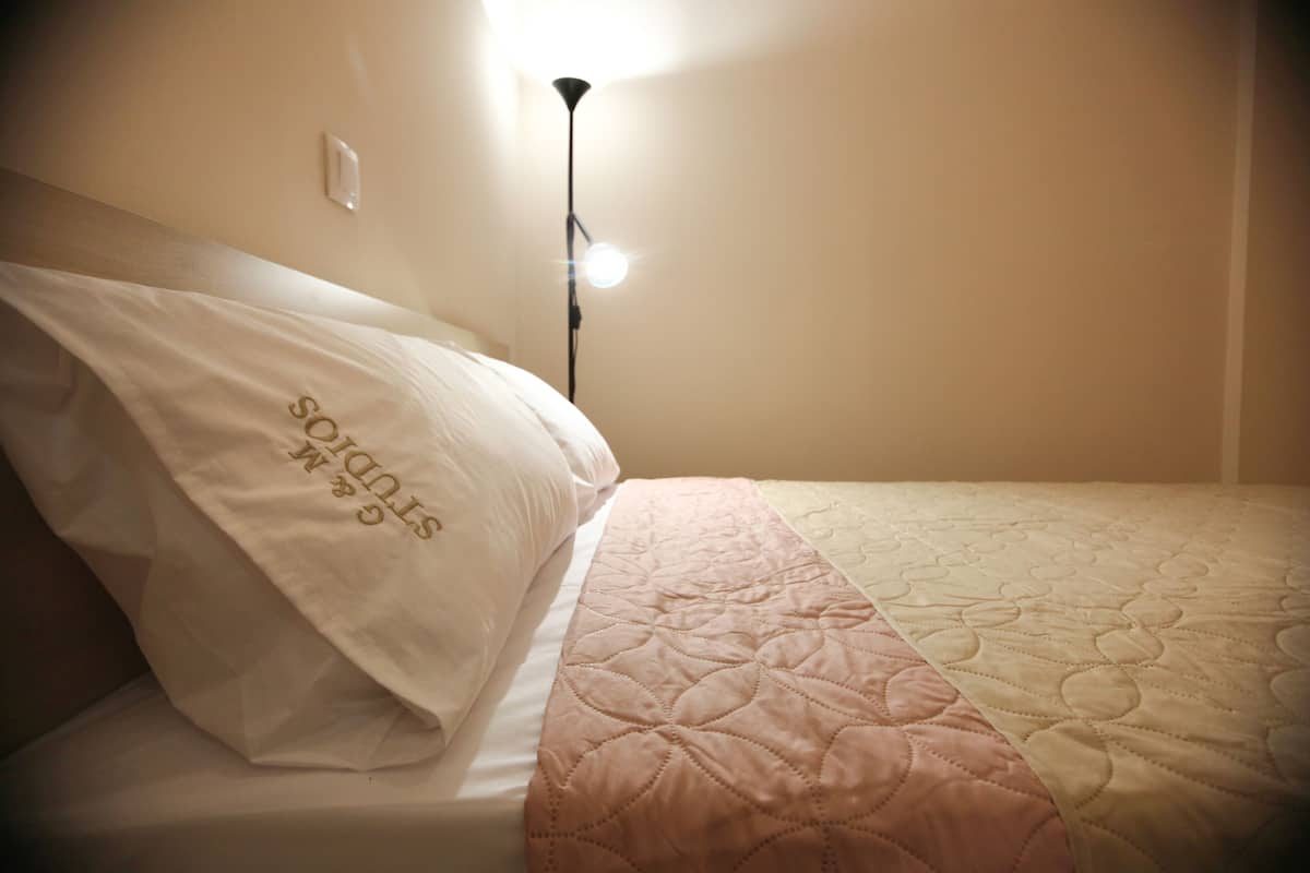 A bed with a white pillow and a pink quilt, a bedside lamp, and a wall socket.