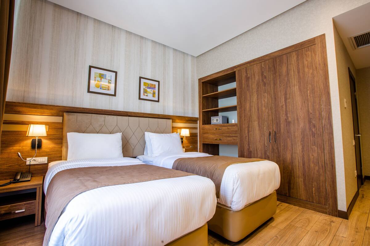 A hotel room with two beds, wooden headboard, bedside lamps, a wardrobe, and a small desk with a telephone.