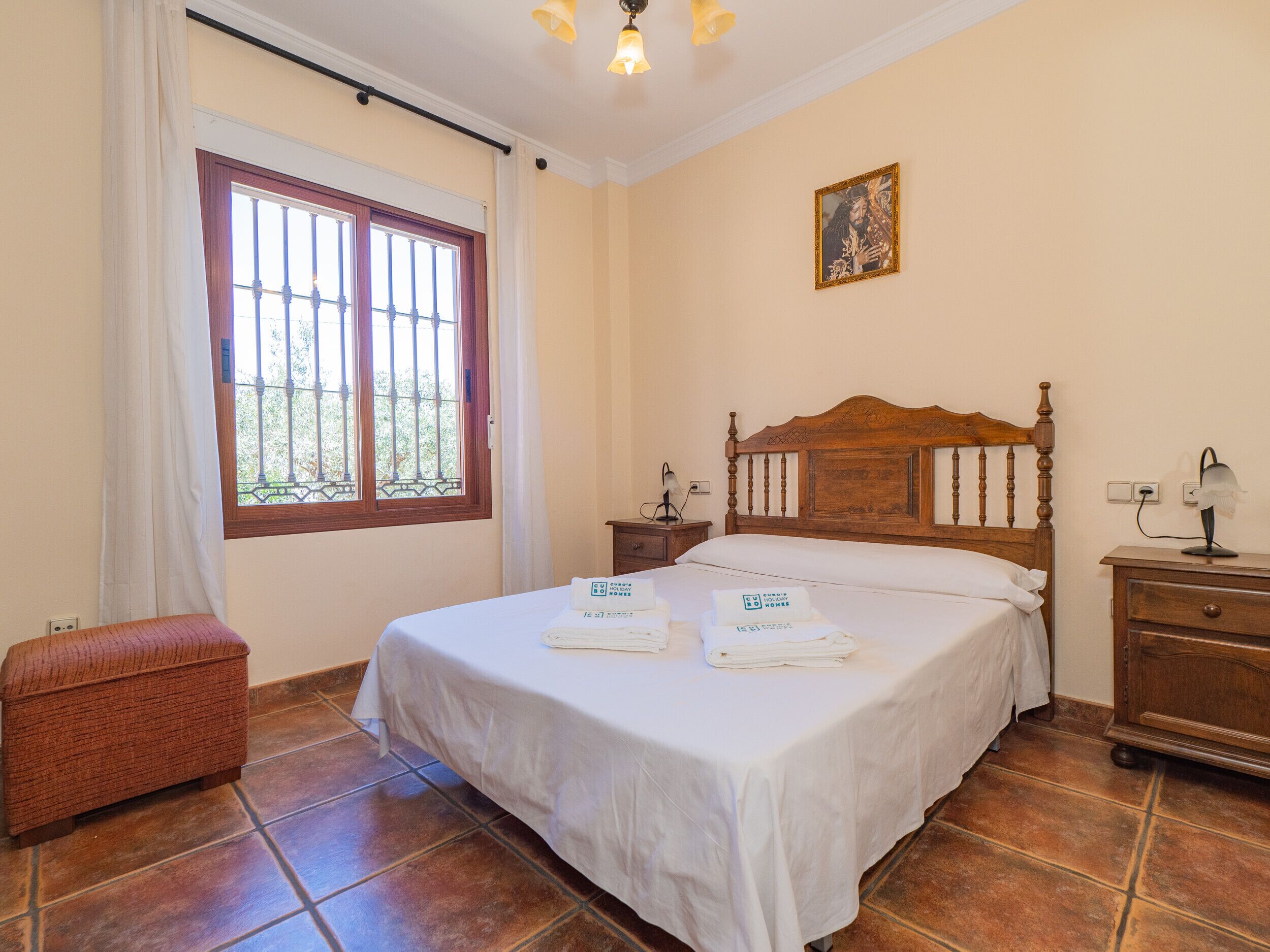 A neatly arranged bedroom with a large bed, a bedside table, a nightstand, a bench, and a window with bars.