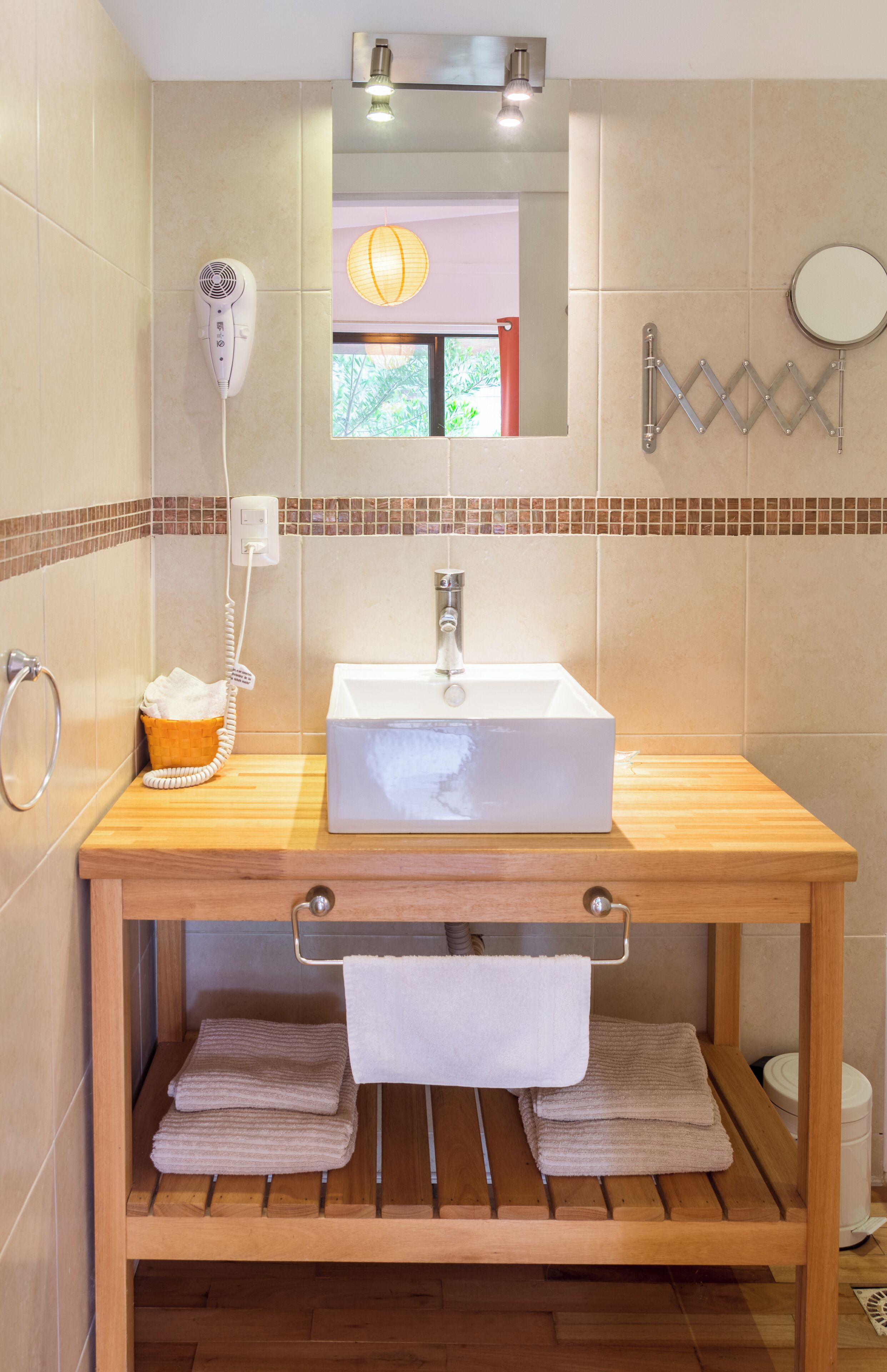 Un bagno con un mobiletto contenitore in legno, un lavandino bianco, un porta asciugamani, un asciugacapelli e uno specchio.