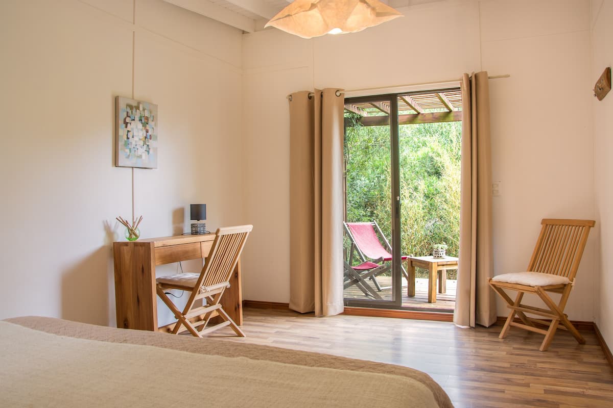Ein Schlafzimmer mit einem Bett, einem Schreibtisch mit Stuhl, einem Fenster mit Vorhängen und Blick auf einen Außenbereich mit Tisch und Stühlen.