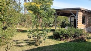 Un chalet rustique en rondins de bois, doté d’un porche abrité, entouré d’un jardin luxuriant où poussent différentes espèces d’arbustes et d’arbres.
