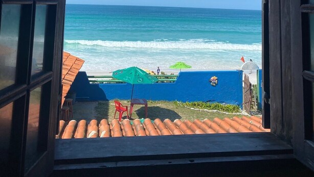 Een open raam met uitzicht op de oceaan en een strand met parasols en een persoon.