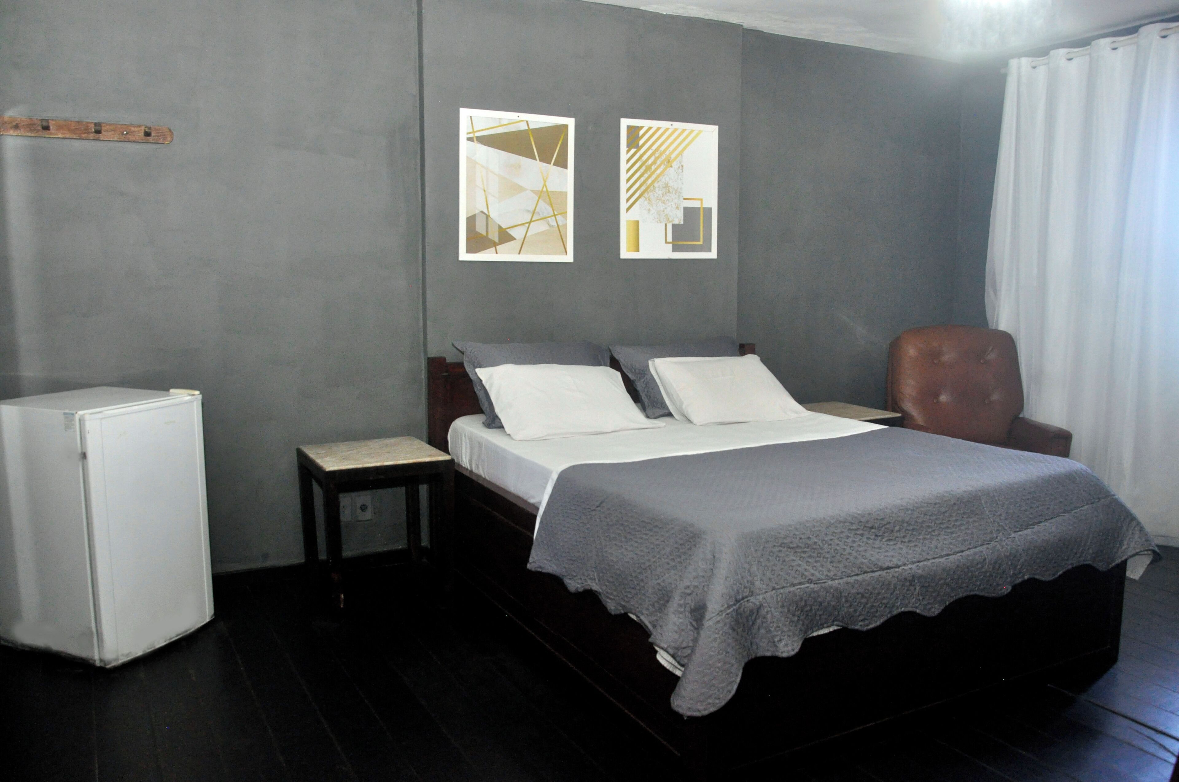 Un dormitorio con cama, una silla, una mesita de noche y un electrodoméstico pequeño y blanco.