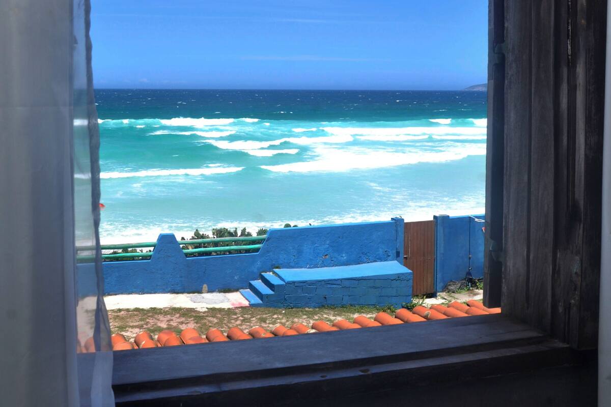 A view through an arched window of the ocean, a wooden structure, and a blue wall with steps leading down to the beach.