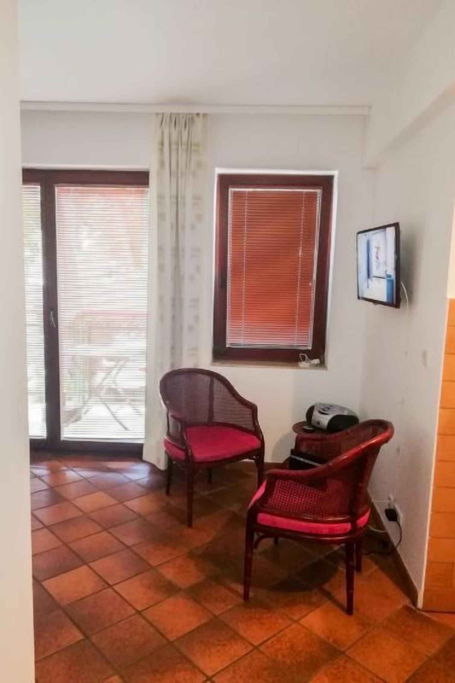 Une pièce avec deux fauteuils en osier, une fenêtre avec des stores, une porte donnant sur l’extérieur et un sol carrelé.