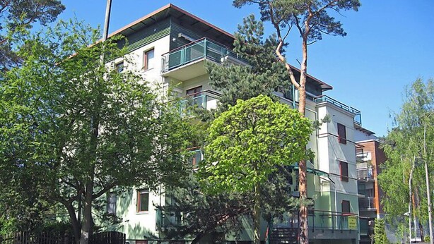 Un edificio residencial de varios pisos con balcones, rodeado de árboles y una entrada con portón.