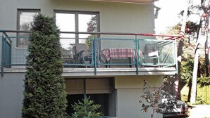 Un edificio de dos plantas con un balcón, un coche aparcado en la planta baja y un seto frondoso al frente.