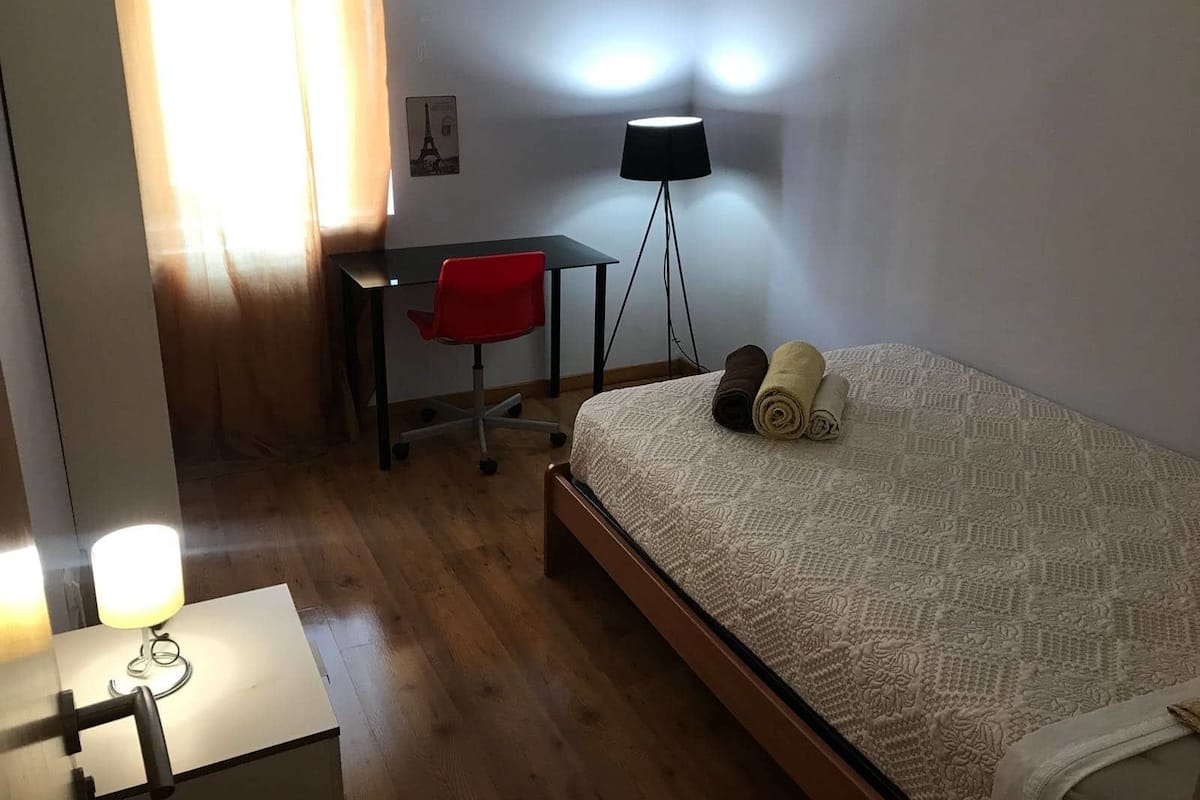 Makuuhuoneessa on sänky, työpöytä, tuoli, lamppu ja ikkuna, jossa on verhot.