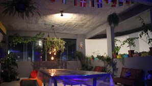 A cozy indoor space with a table and chairs, surrounded by potted plants and a hanging plant. Flags are visible on the ceiling.