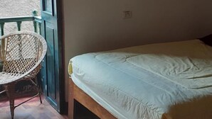 Une chambre avec un plancher en bois, un lit avec un matelas blanc, une chaise en osier et une fenêtre donnant sur un mur extérieur.