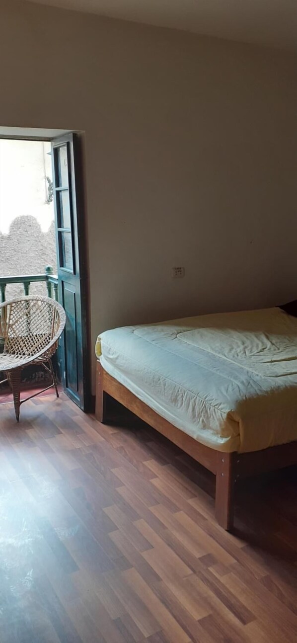 A bedroom with a wooden floor, a bed with a white mattress, a wicker chair, and a window with a view of a wall outside. - The Grasshopper Hostels AV Alta (Cusco)