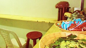 Une pièce avec une table en bois, un panier en osier rempli de feuilles, un tabouret rouge, une chaise en plastique et un mur orné d’une série de petites images.