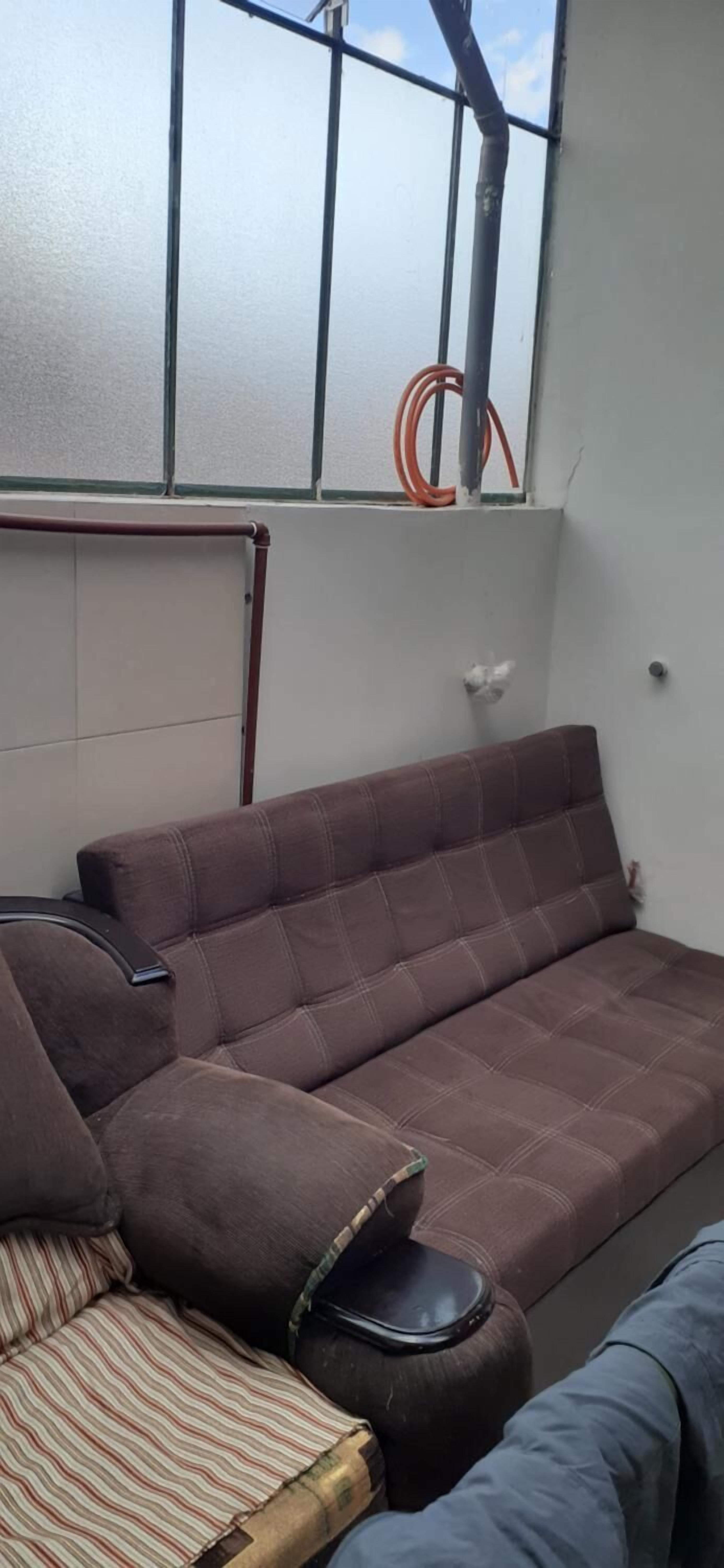 A brown sofa with a striped cushion, a round ottoman, and a window with a view of the sky and a red hose.